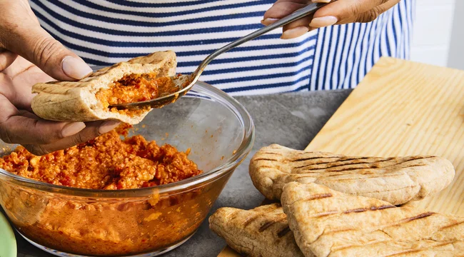 Person in a striped apron standing behind a grey table filling a pita pocket with romesco sauce over a glass bowl of romesco sauce