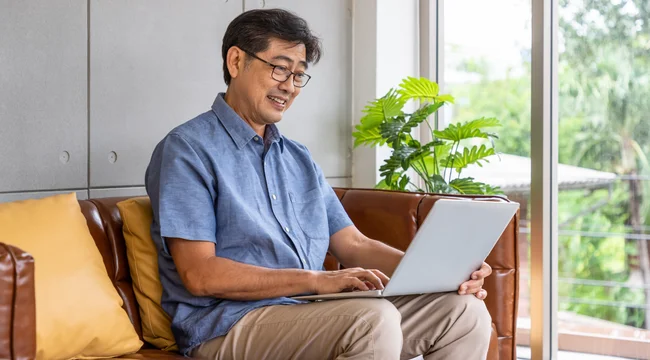 A man in a blue shirt on his computer while sitting on a brown couch