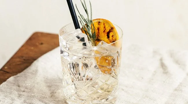 Glass of orange tonic with a sprig of rosemary and burnt orange garnish placed on a white table cloth