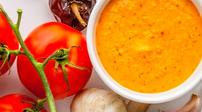 Bowl of romesco sauce surrounded by fresh tomatoes, garlic cloves, and roasted bell peppers.